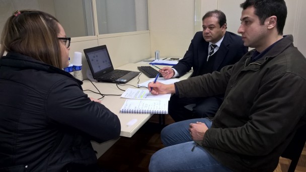 Em primeiro plano, dois servidores do Daer sentados em frente a uma mesa com um notebook, sendo uma moça de costas para a foto e um rapaz de lado. Ao fundo, um consultor, de terno e gravata e também sentado, escuta atento ao que a moça fala.