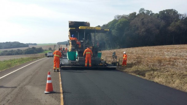 A imagem é de uma obras em uma rodovia, com uma equipe de operários vestidos com macacão laranja, acompanhando uma máquina que está colocando asfalto na via.
