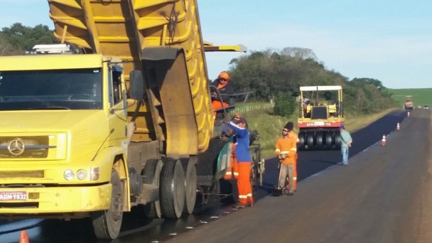A fot mostra um caminhão de obra e um rolo compressor logo atrás, numa rodovia em trecho reto sendo recapeada. Seis funcionários estão junto ao caminhão, realizando obras.