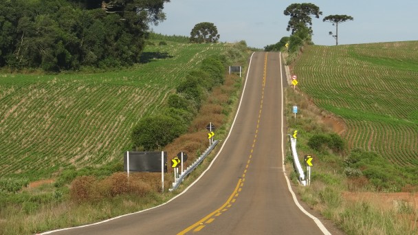 A foto mostra um trecho em reta, elevado, do acesso municipal pavimentado do município de Água Santa. Às margens, lavouras e árvores nativas.
