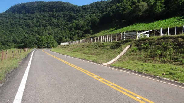 A foto mostra uma rodovia pavimentada e com pintura de sinalização demarcando as duas pistas. O trecho é em reta, com montanhas ao fundo.