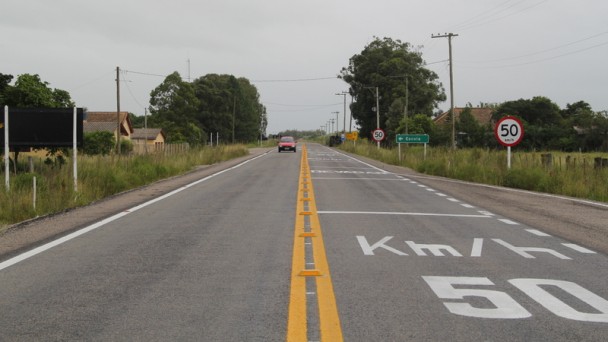 A foto mostra uma rodovia pavimentada, com pintura na pista indicando o limite de velocidade de 50 quilômetros por hora, com placas de trânsito às margens