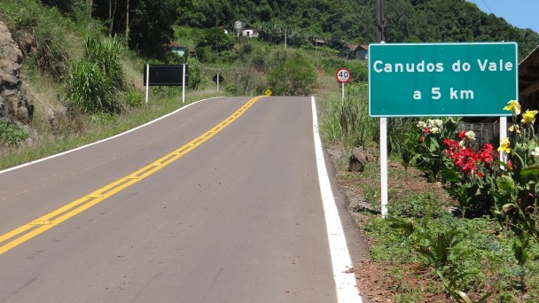 A foto mostra uma estrada pavimentada, com tachões e pintura dividindo o acostamento e as duas pistas. Às margens, uma placa dizendo que Canudos do Vale está a 5 quilômetros de distância e flores coloridas ao redor.
