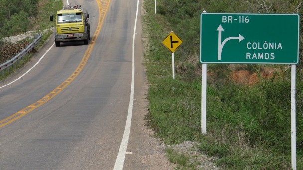 A imagem mostra uma rodovia pavimentada, com tachões e sinalização de trânsito, com destaque para uma placa indicando a localidade de Colônia Ramos e a BR-116. Um caminhão trafega em uma das pistas.