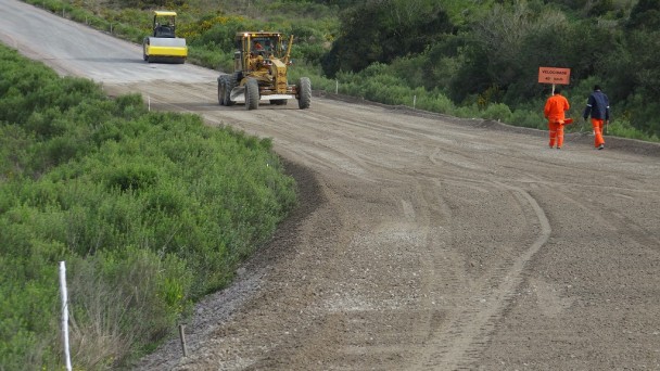 A foto mostra obras de pavimentação na rodovia de acesso ao município de Pedras Altas. Na pista, um rolo compressor, uma patrola e dois operários a pé.
