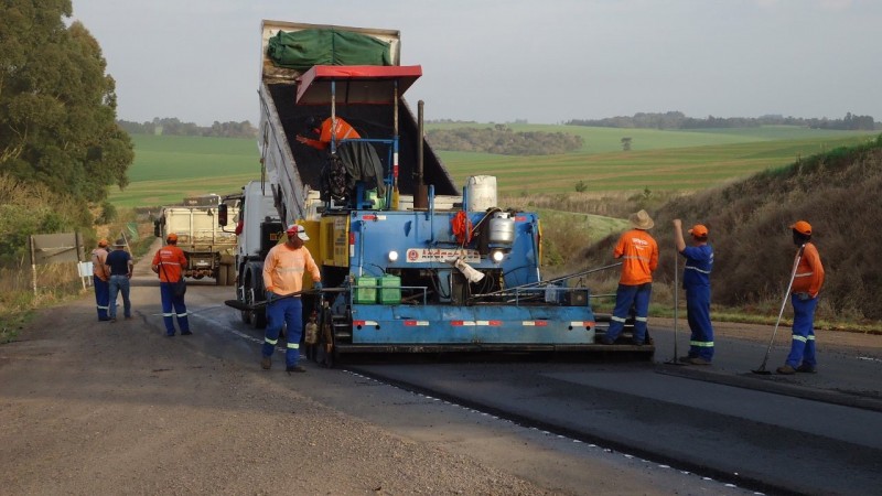 Na foto, operários fazem obra de recapeamento de uma rodovia pavimentada, com o uso de máquinas e caminhões.