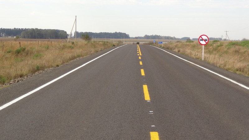 A foto apresenta uma pista pavimentada e com sinalização horizontal e vertical. Nas margens, vegetação baixa.